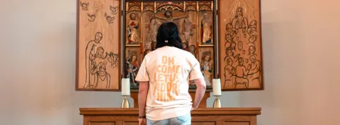 Rückenansicht des „Adore Him“-Shirts mit dem Aufdruck „Oh come let us adore him“ vor einem Altar